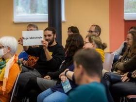 Menschen sitzen bei einem Fachtag im Plenaum. Eine Person hält ein Schild nach oben auf dem "Jugendstrategie" steht.