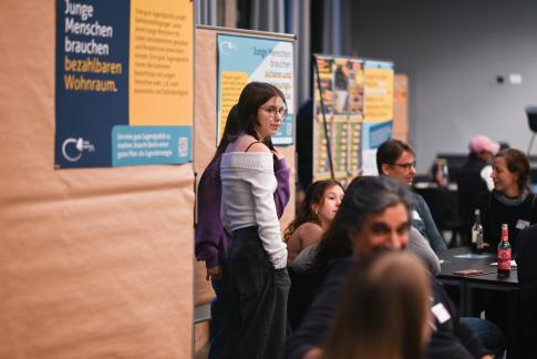 Eine junge Frau steht vor Plakaten zu Jugendthemen, umgeben von Menschen in einer Diskussionsrunde bei einer Veranstaltung.